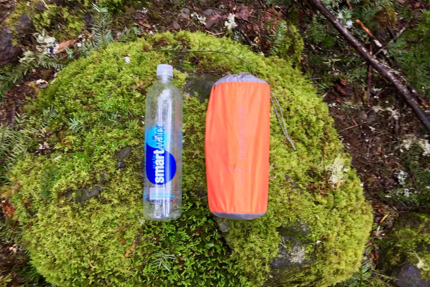 a sea to summit ultralight insulated sleeping pad packed in its stuff sack and placed on a mossy rock next to a one liter smart water bottle to show the small packed size of the pad