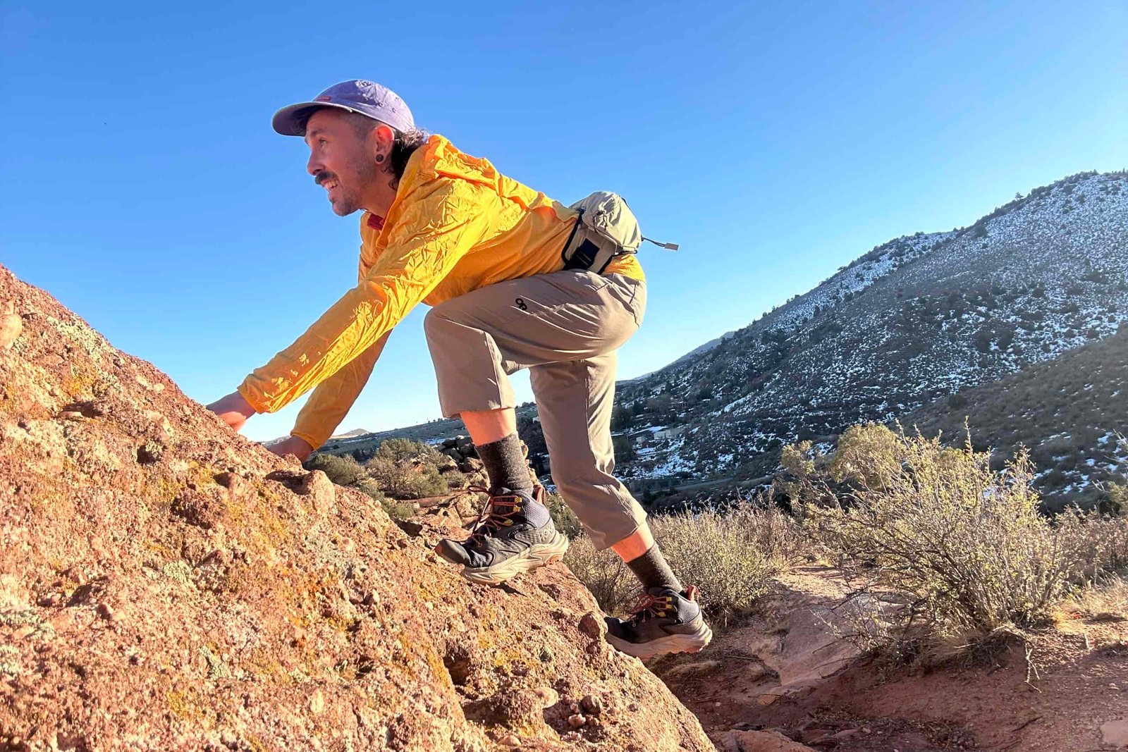A man scrambles up some rocks in a cap and yellow jacket.
