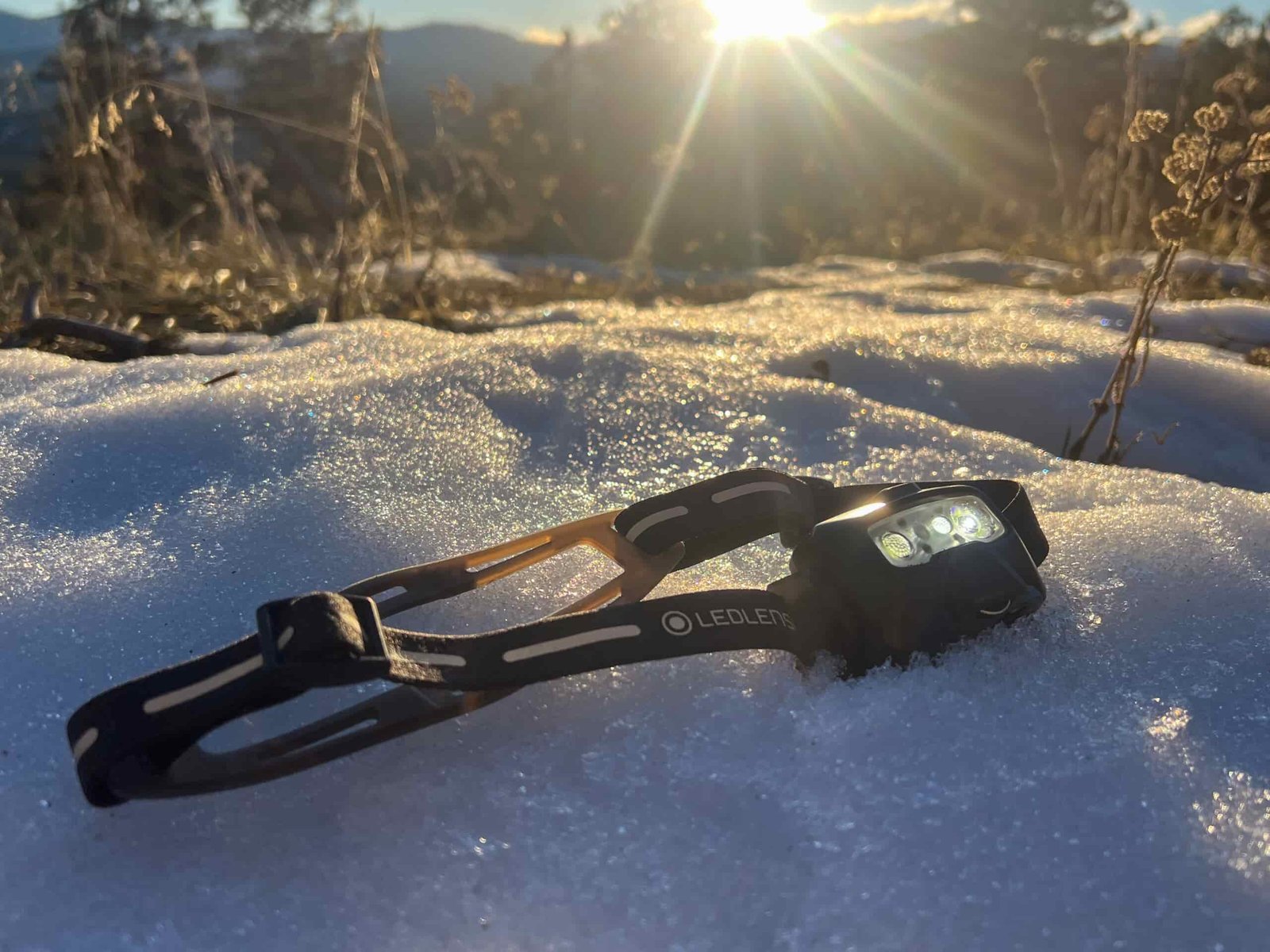 The image shows the Ledlenser HF6R Signature headlamp resting on snow, illuminated by the golden glow of the sun in the background. The headlamp is positioned at an angle to show its front-facing lenses, and its strap, marked with the Ledlenser logo, is visible. The snow beneath the headlamp is sparkling in the sunlight, and the surrounding landscape includes distant mountains and a few plants.