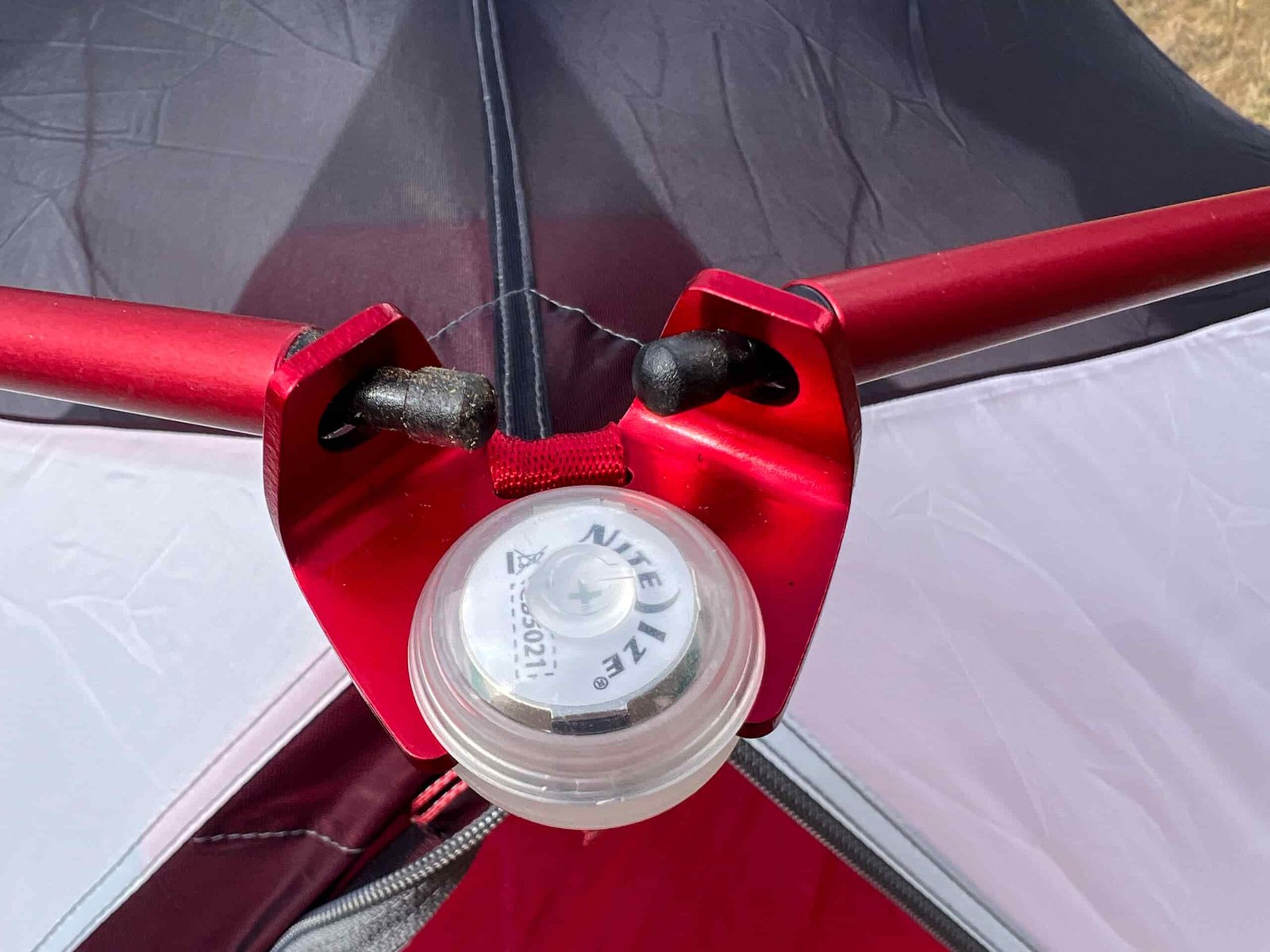 Close-up of a red aluminum pole and attachment point on a tent, with a small round porch light mounted at the junction. The tent fabric is a mix of red and white, with a mesh window visible.