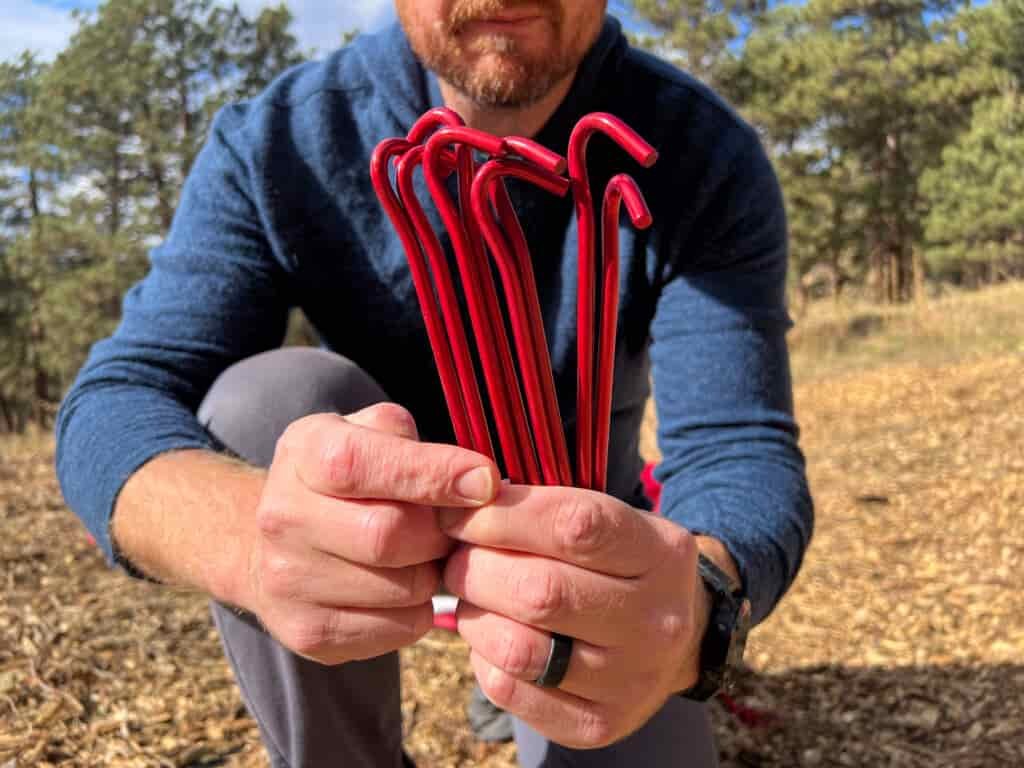 A person holding several red tent stakes in their hands, with the stakes visibly bent in a "hook" shape. The background shows a wooded area with scattered dry leaves.