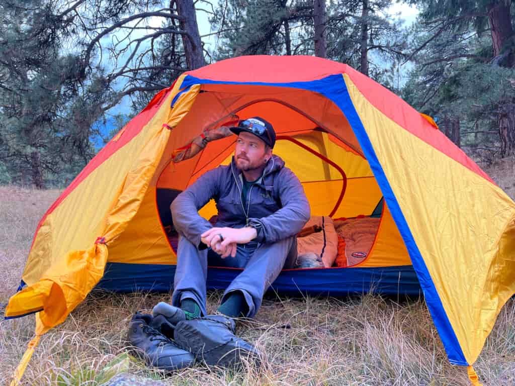 A camper sits cross-legged in the entrance of a brightly colored tent, showcasing its low headroom and cozy interior. The vibrant orange, yellow, and blue tent is pitched in a wooded area, with a pair of hiking boots placed on the grassy ground outside.