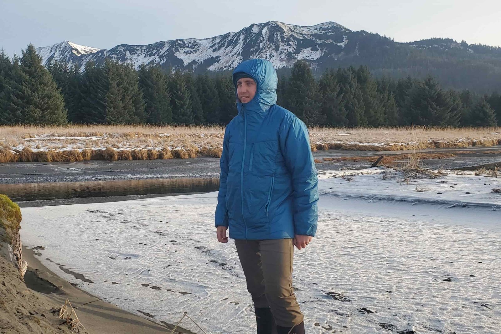 A man stands with a hooded jacket in front of a mountain view.