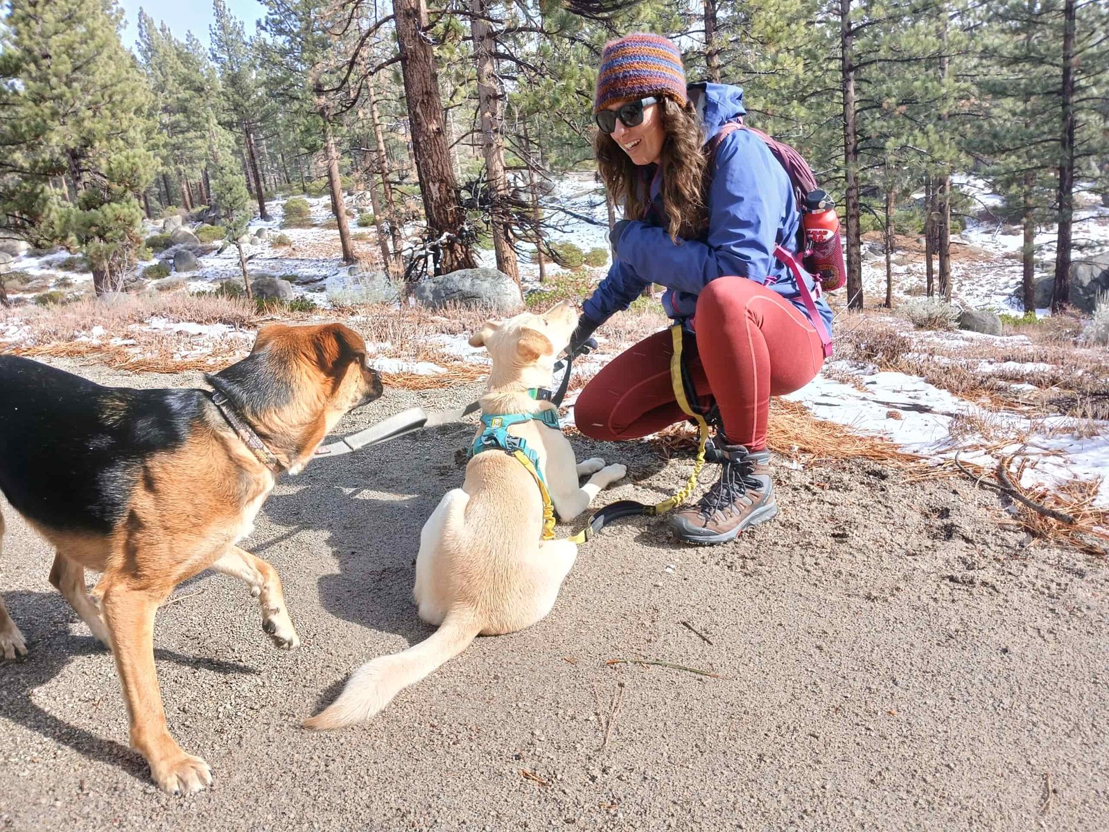 Woman hiking in reddish/orange leggings on a dirt trail with a little snow on the sides of the trail. She's wearing a small purple backpack, a different color purple rain jacket, and a beanie, plus brown and grey hiking boots. There are pines trees, bushes, and granite rocks off to the sides of the trail.