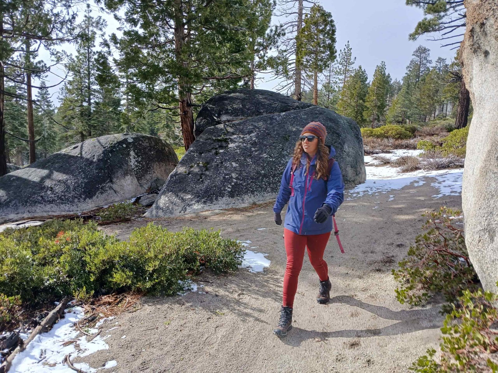 Woman hiking in reddish/orange leggings on a dirt trail with a little snow on the sides of the trail. She's wearing a small purple backpack, a different color purple rain jacket, and a beanie, plus brown and grey hiking boots. There are pines trees, bushes, and granite rocks off to the sides of the trail.