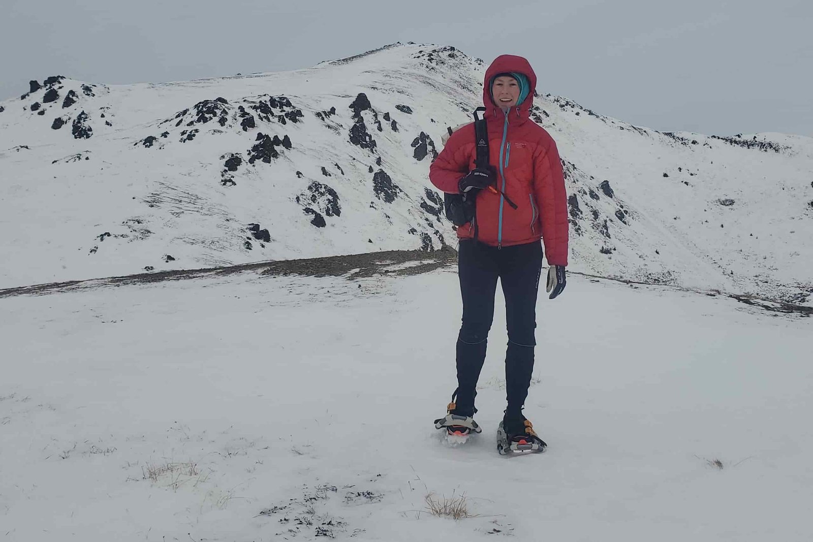 A woman walks along a snowy ridge with a mountain in the background.