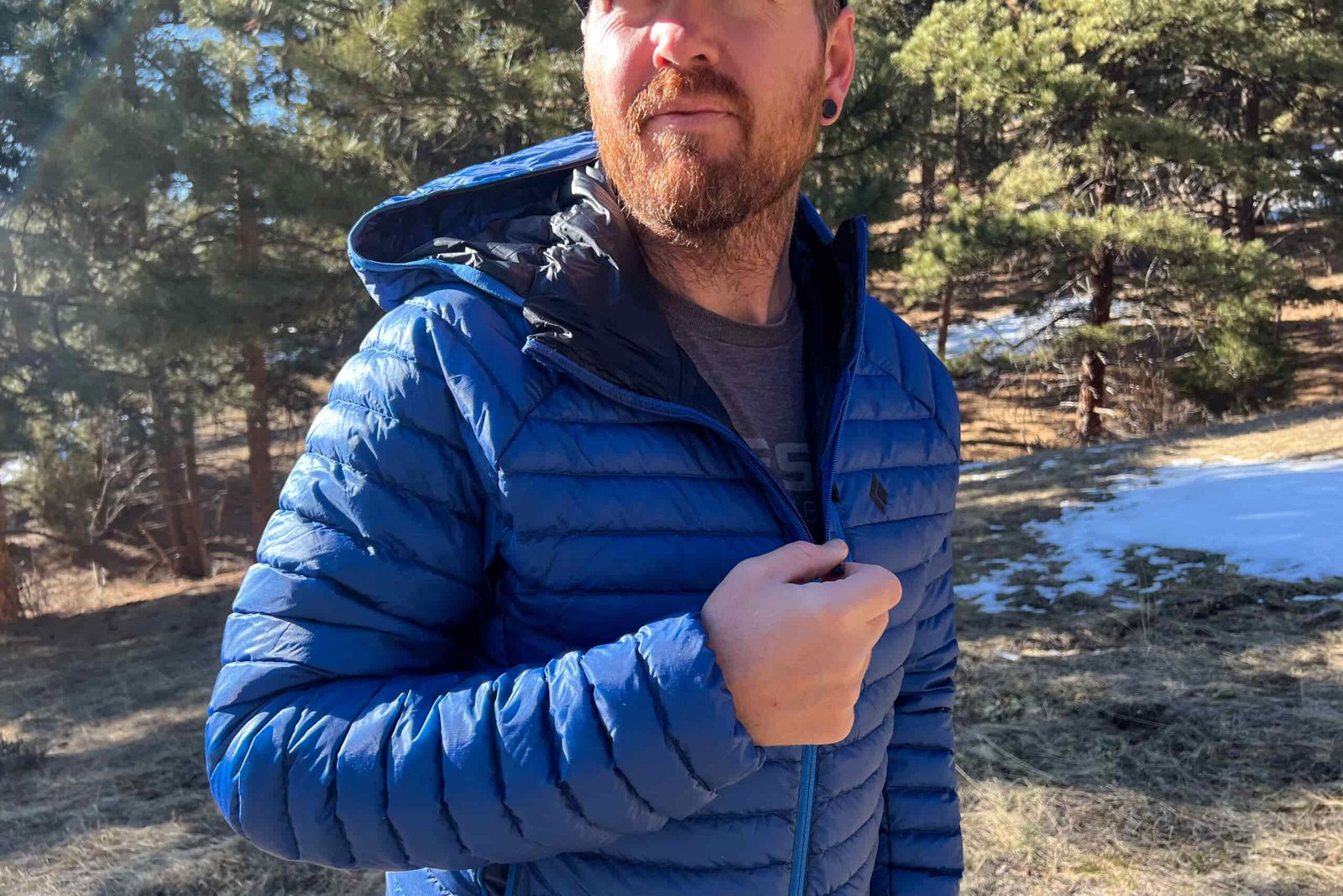 A person adjusting the zipper of the Black Diamond Approach Down Hoody, standing in a sunlit forest with light snow on the ground.