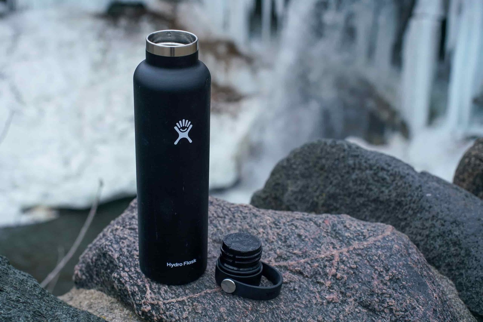 The Hydro Flask Standard Mouth, with its lid off, sits on a rock with a partially frozen waterfall in the background.