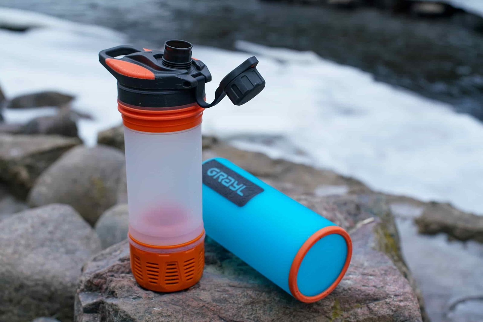 The Grayl GeoPress water bottle's inner press and outer cup sit separately on a rock with a partially frozen stream behind it.