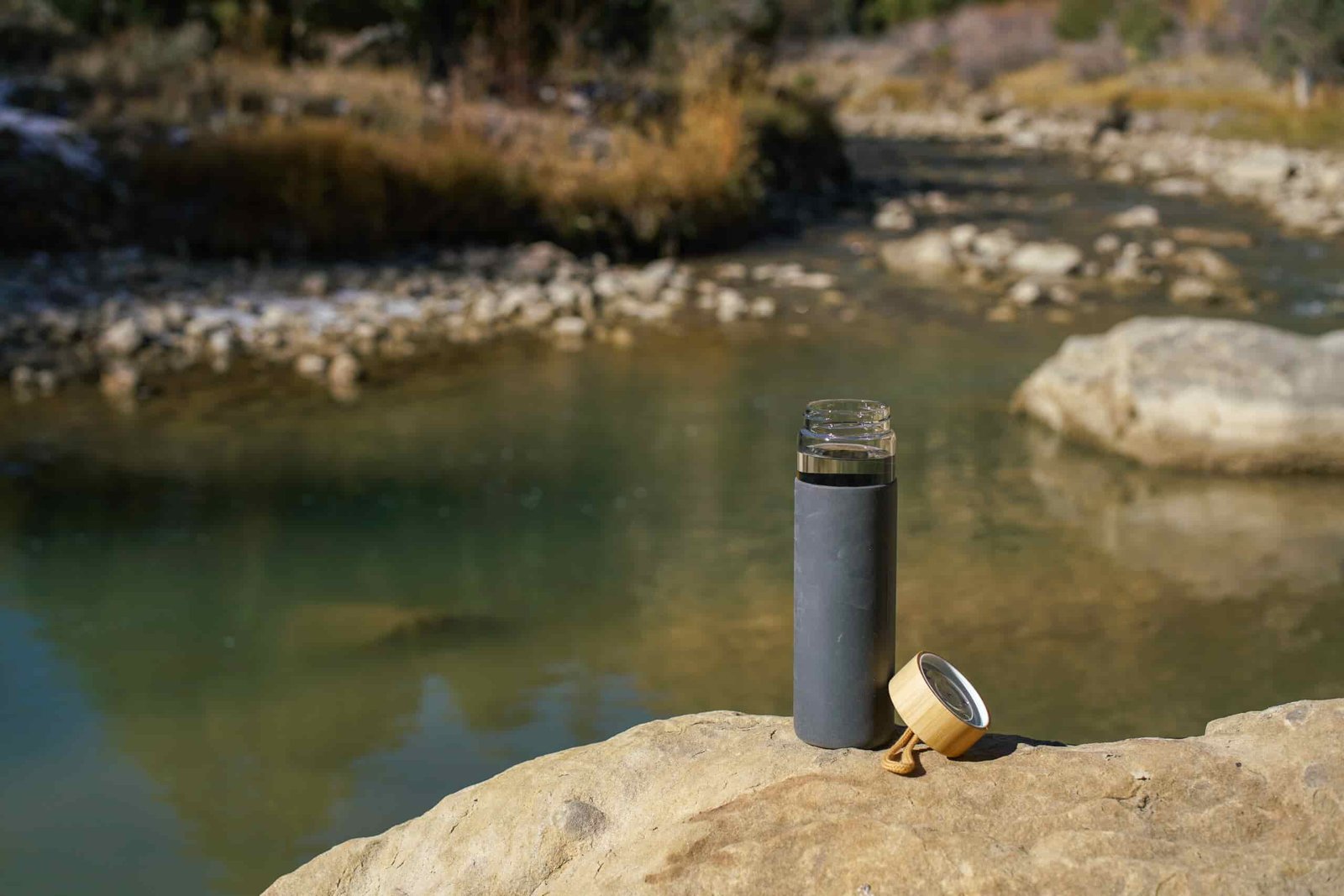 The Yomious Glass water bottle with its bamboo lid beside it, sits on a rock with a river in the background.