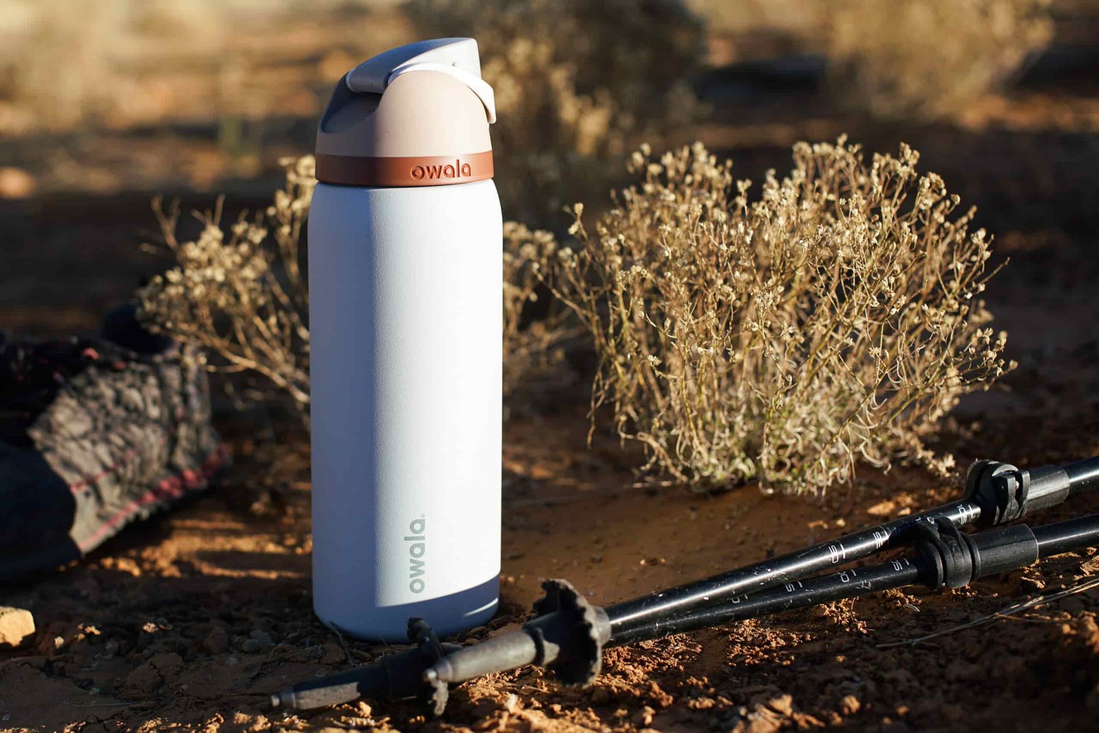 The Owala FreeSip Vacuum Insulated water bottle sits on the ground with trekking poles beside it.