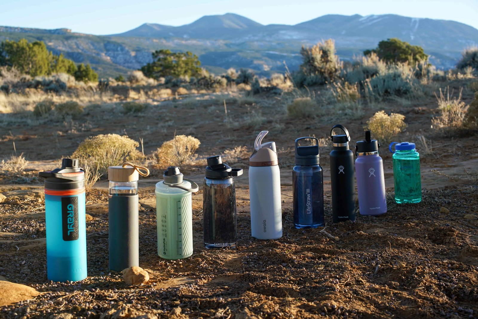 The Grayl GeoPress, Yomious Glass, Hydrapak Flux, CamelBak Chute Mag Renew, Owala FreeSip Vacuum Insulated, LifeStraw Go Series, Hydro Flask Standard Mouth, Hydro Flask Wide Mouth, Nalgene Wide Mouth sit on the ground in a line with mountains in the background.