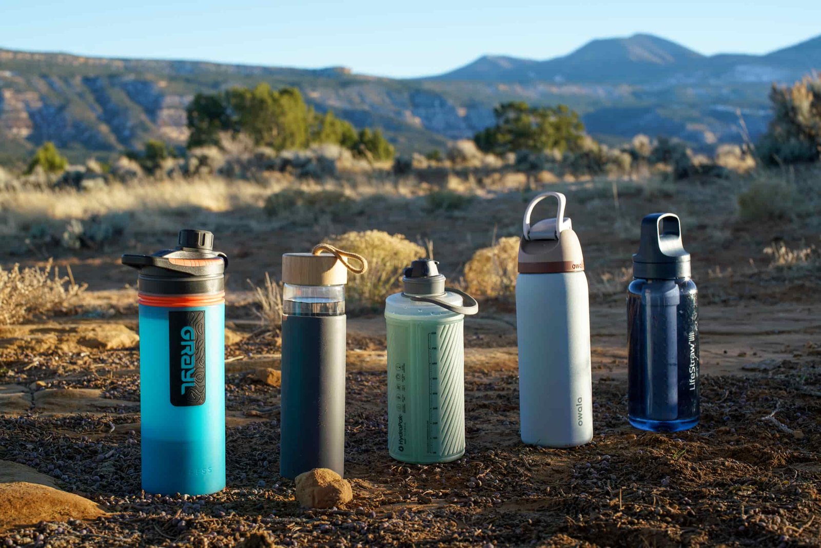 Grayl GeoPress, Yomious Glass, Hydrapak Flux, Owala FreeSip Vacuum Insulated, and LifeStraw Go Series water bottles sit on the ground in a line with mountains in the background.