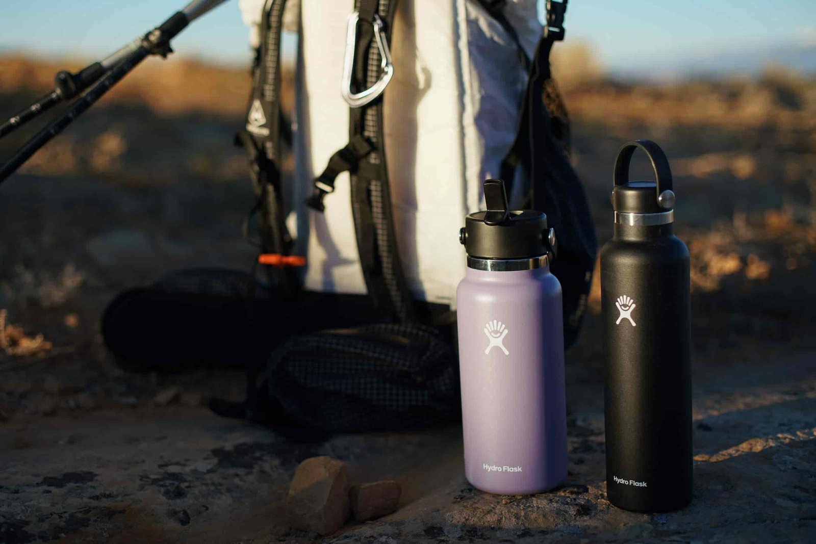 The Hydro Flask Wide Mouth and Hydro Flask Standard Mouth sit next to each other on the ground with a backpack behind them.
