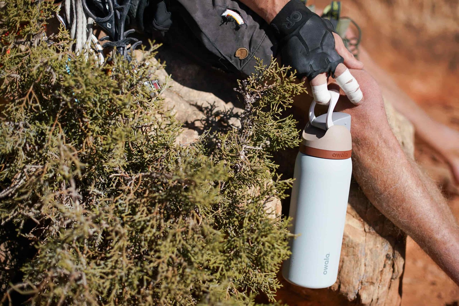 A person wearing crack climbing gloves and with taped fingers holds the Owala FreeSip Vacuum Insulated water bottle by its carrying handle.
