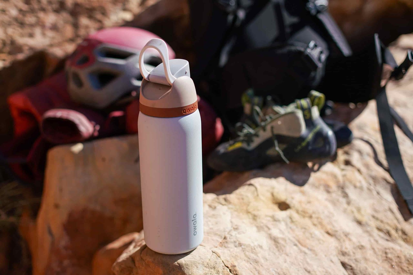 The Owala FreeSip Vacuum Insulated water bottle sits on a rock with climbing shoes and helmet in the background.