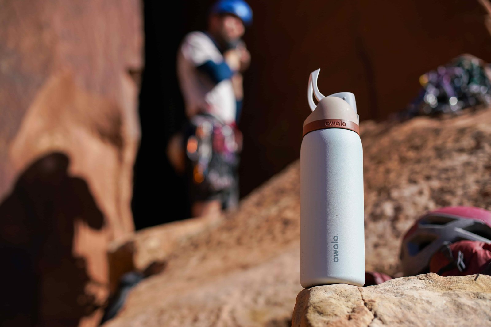 The Owala FreeSip Vacuum Insulated water bottle sits on a rock with a climber in the background.