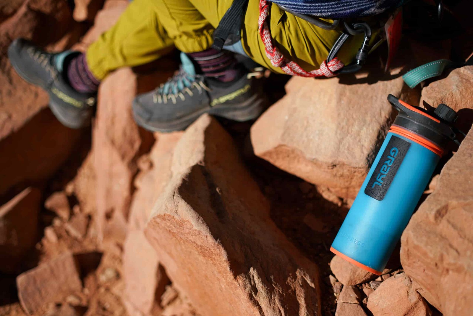 The Grayl GeoPress water bottle leans against a rock with a person wearing a climbing harness sitting partially visible beside it.