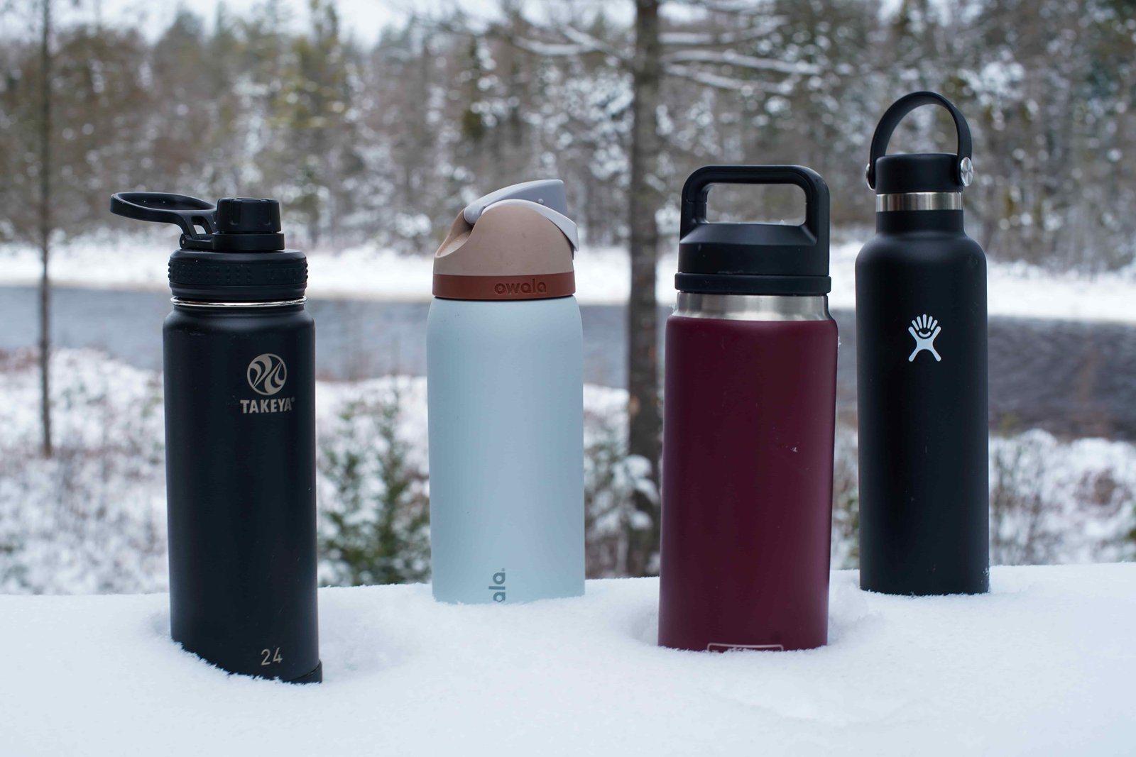 The Takeya Actives Insulated, Owala FreeSip Vacuum Insulated, YETI Rambler, and Hydro Flask Standard Mouth water bottles sit in the snow with a snowy lake in the background.