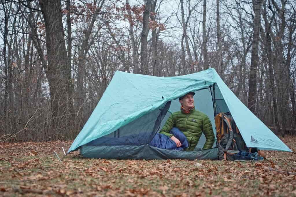 a man in a green coat sits in a one person tent in a forest clearing