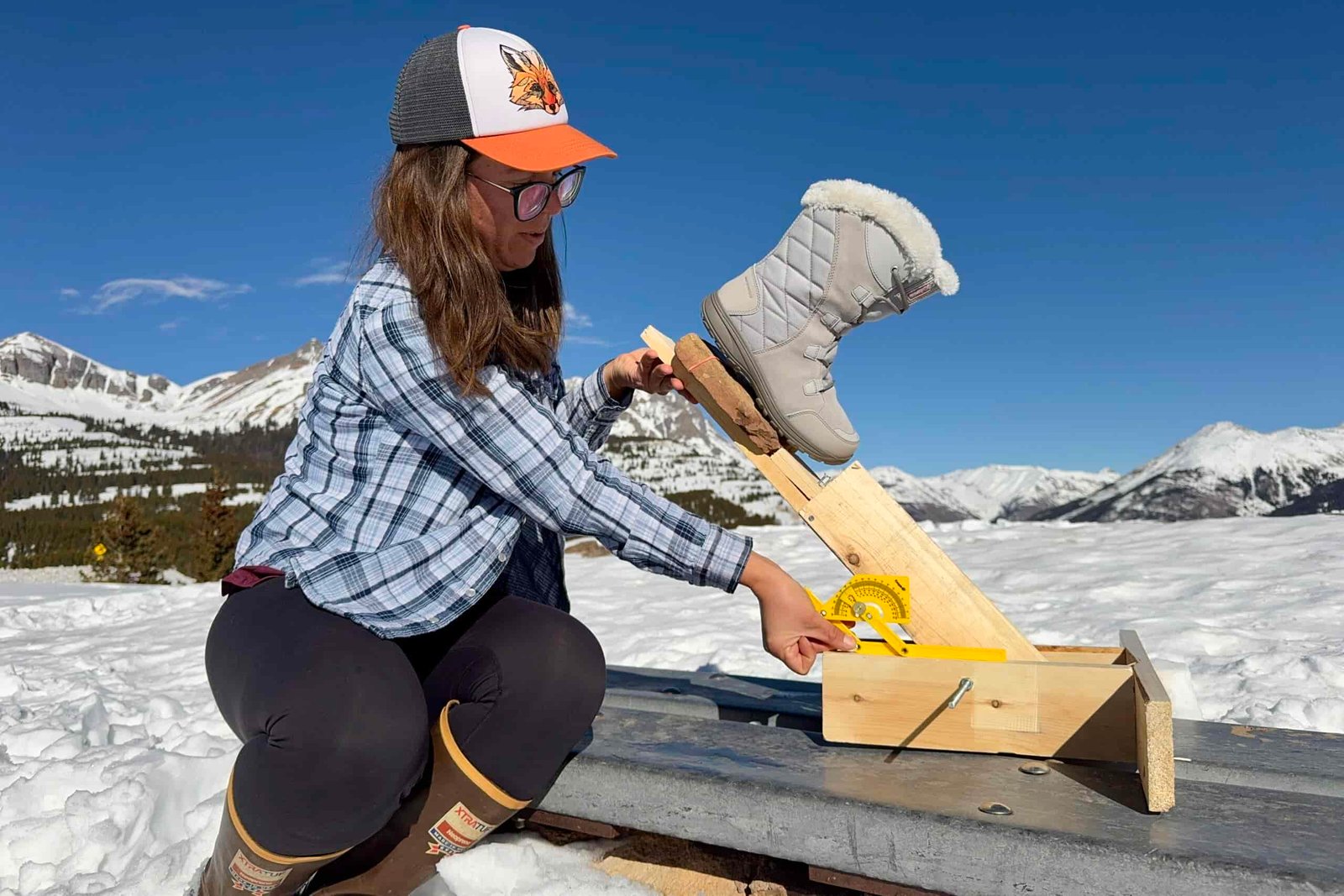 A person is measuring the traction of the Ice Maiden boot. The boot is sitting on a rock that is perched on a wooden lever arm and the person is holding a protractor up to the device. The measurement is taking place in the snow in the mountain.