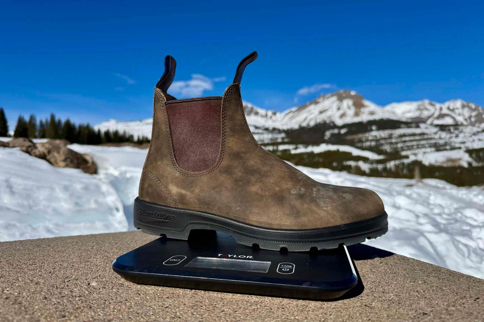 A close up of the Blundstone boot sitting on a kitchen scale. The scale is sitting on a concrete wall in the mountains and there is snow covering the ground.
