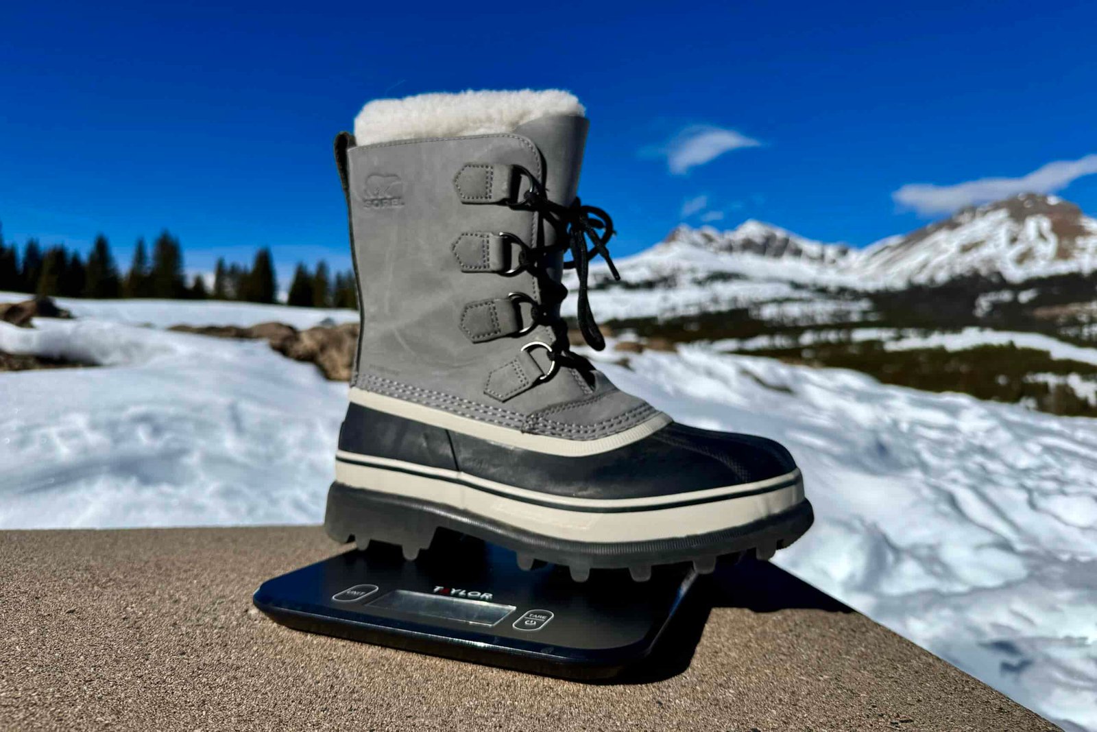 A close up of the Sorel boot sitting on a kitchen scale. The scale is sitting on a concrete wall in the mountains and there is snow covering the ground.