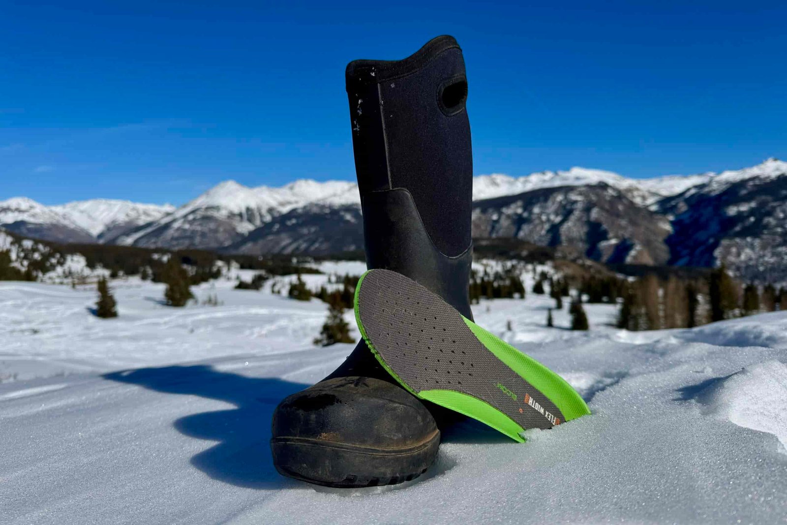 A close up of the Bogs Neo Classic Tall boot with the insole sitting next to it. They are set on some snow in the mountains.