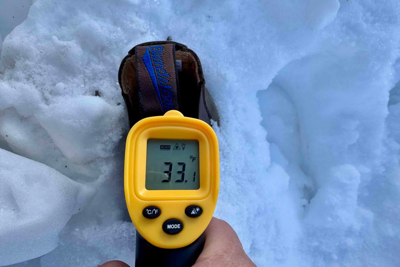 Close up of a hand pointing a laser thermometer into the Blundstone boot which is sitting in the snow.
