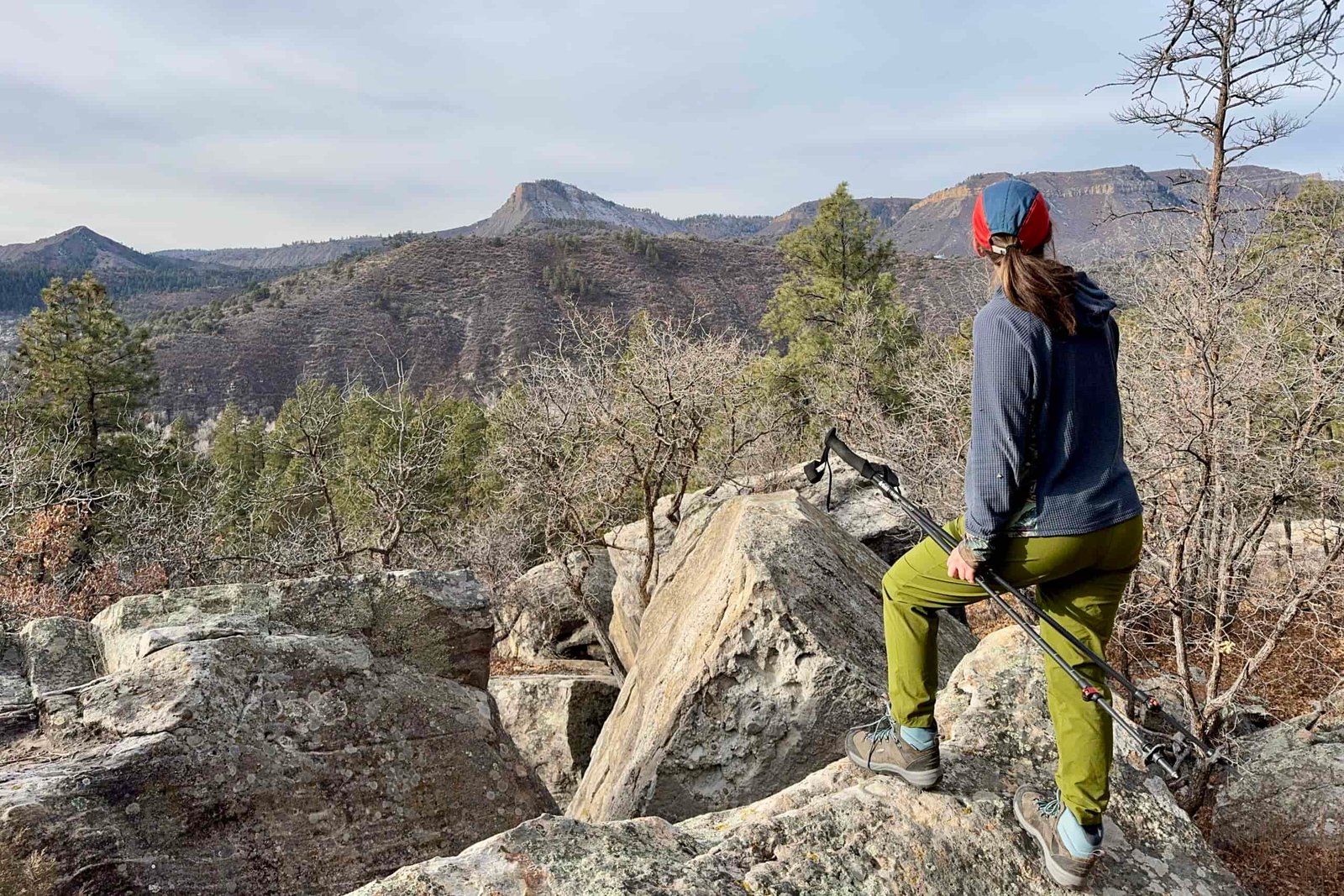 A person is standing on a rock and holding some trekking poles. They are looking out over a mountain view.