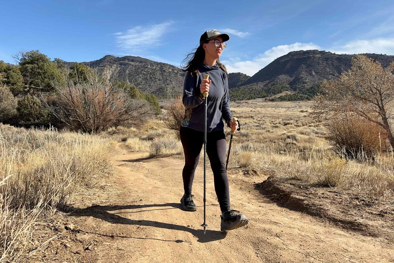 A person is walking down a trail using some trekking poles. They are in desert like area with bushes and pine trees.
