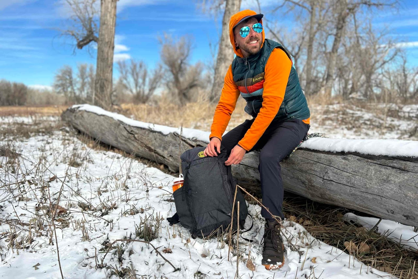 A smiling man sits on a log with his backpack on the ground in a wintry wilderness setting.