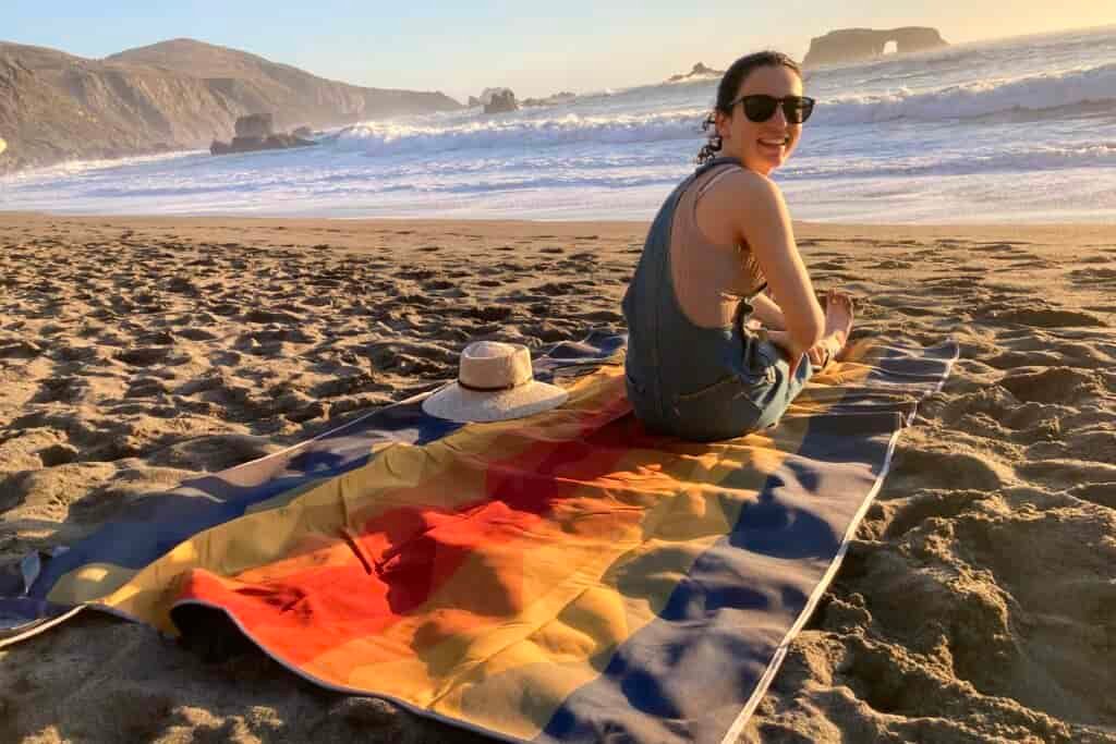 Woman sitting on the Nemo Victory Patio Blanket on the beach, looking over her shoulder at the camera.