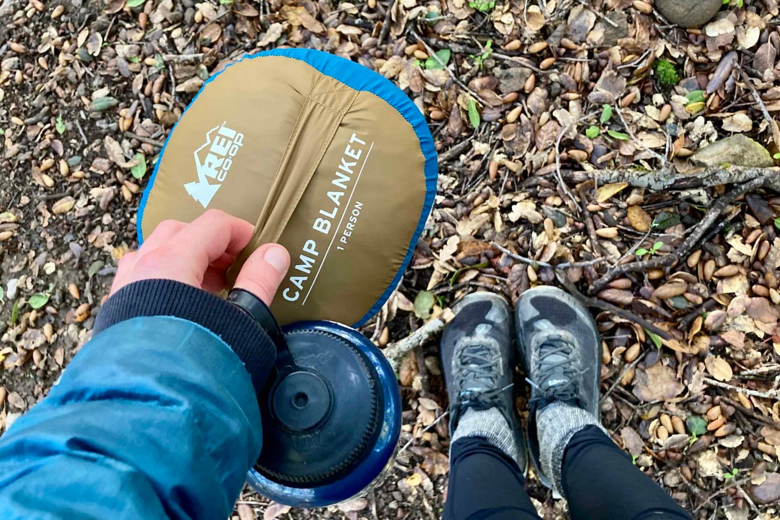 Close up showing someone carrying the REI Camp blanket by the strap on the bottom of the carry bag, while also holding a Nalgene.