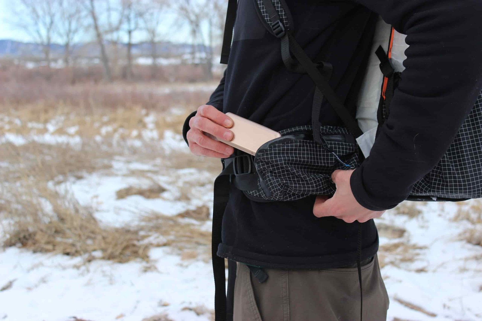 A hiker pulls a battery pack from the hipbelt pocket of a backpack with bare trees and snow in the background.