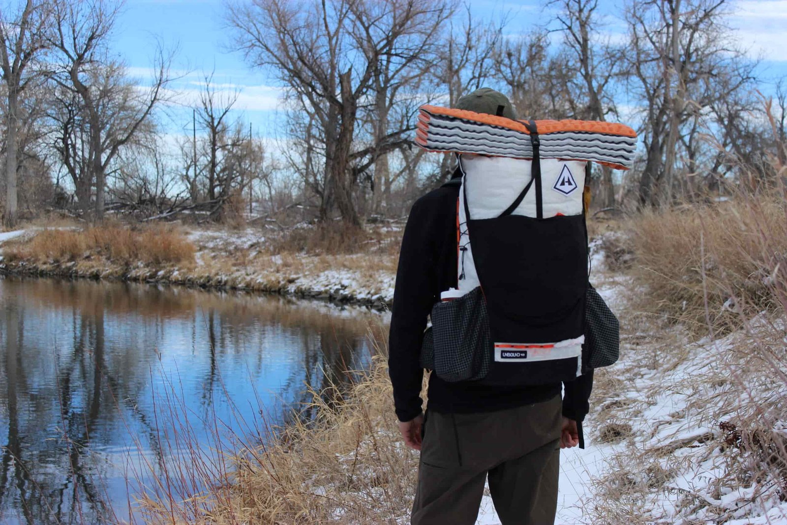 Back view of person wearing backpack against winter stream backdrop. Pack shows contrast between white upper and black lower sections, with folded sleeping pad secured on top. Bare trees and reflection of blue sky in water visible in background.