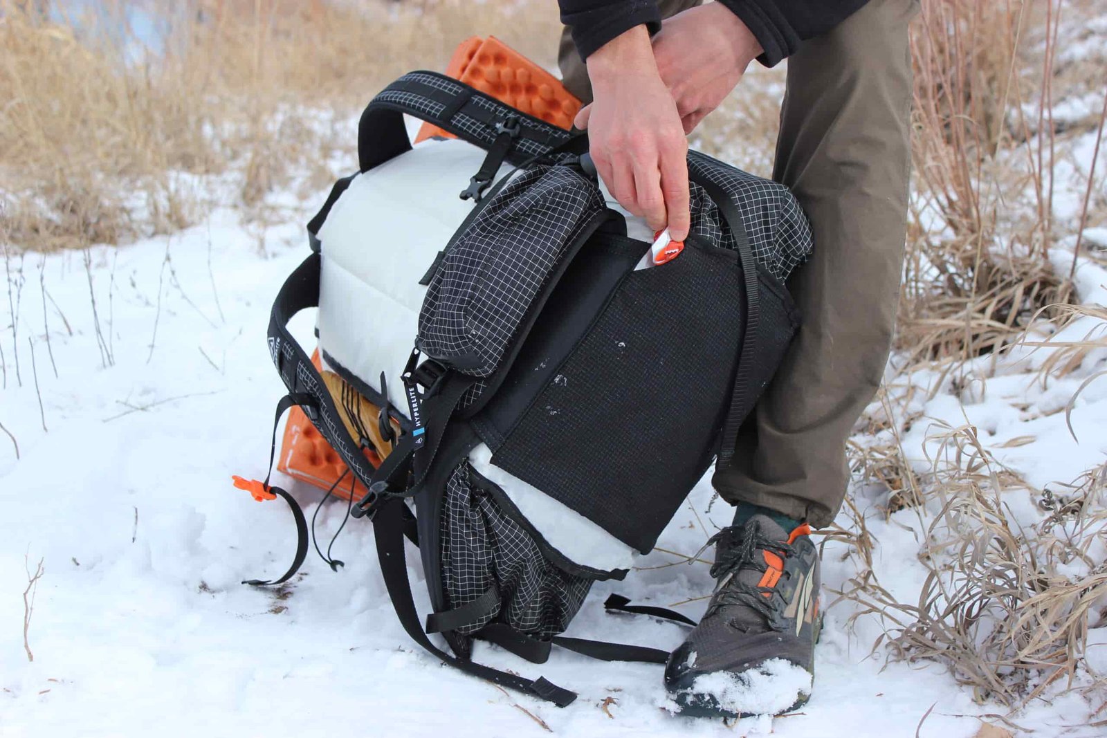 A hand demonstrates accessing the trash port on the bottom of the Unbound 40 pack, with the orange sleeping pad visible in frame. The pack is positioned on snowy ground."