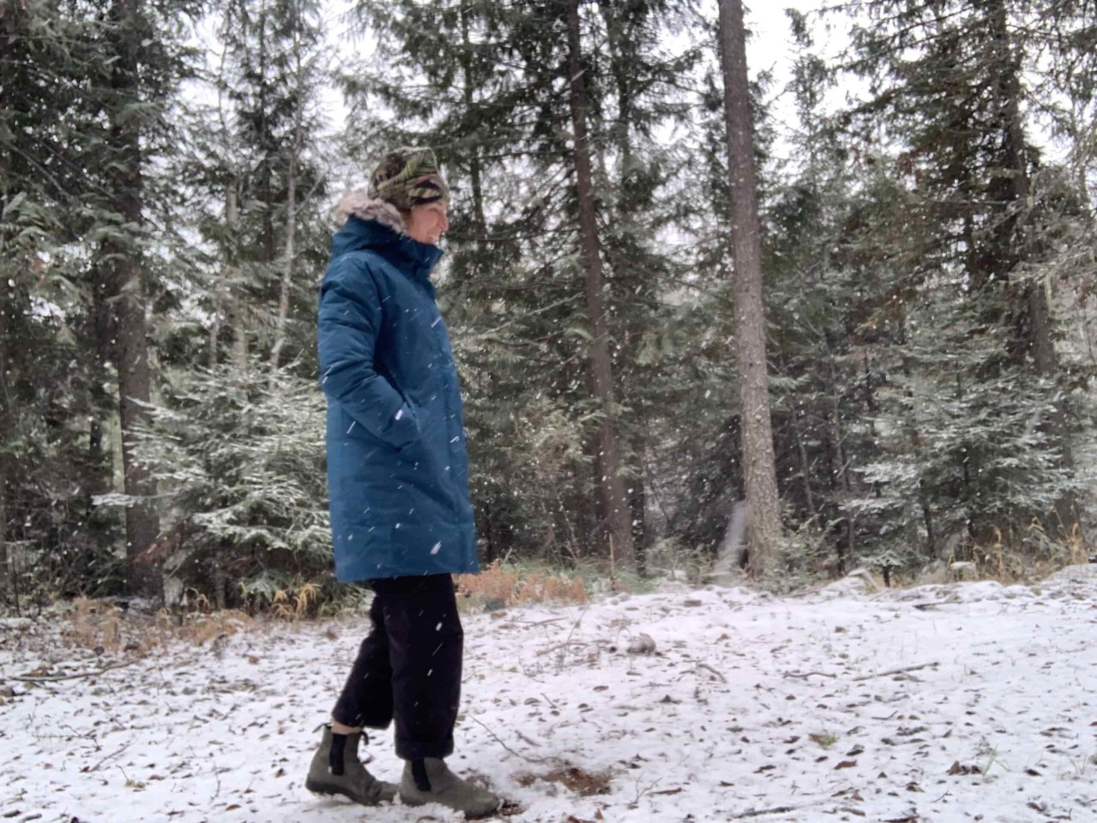 a woman smiling while walking in the wood with light snowfall on the ground. She is wearing a north face arctic parka and has her hands in the pockets