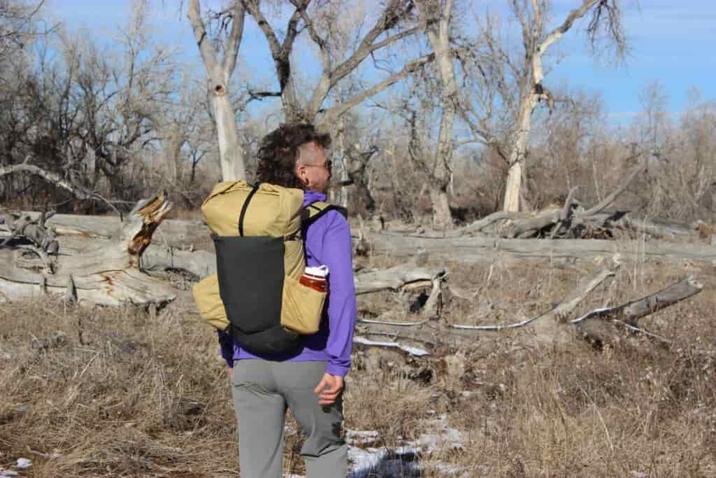A hiker walks away from the camera wearing the pa'lante v2 backpack with trees and blue sky in the background.