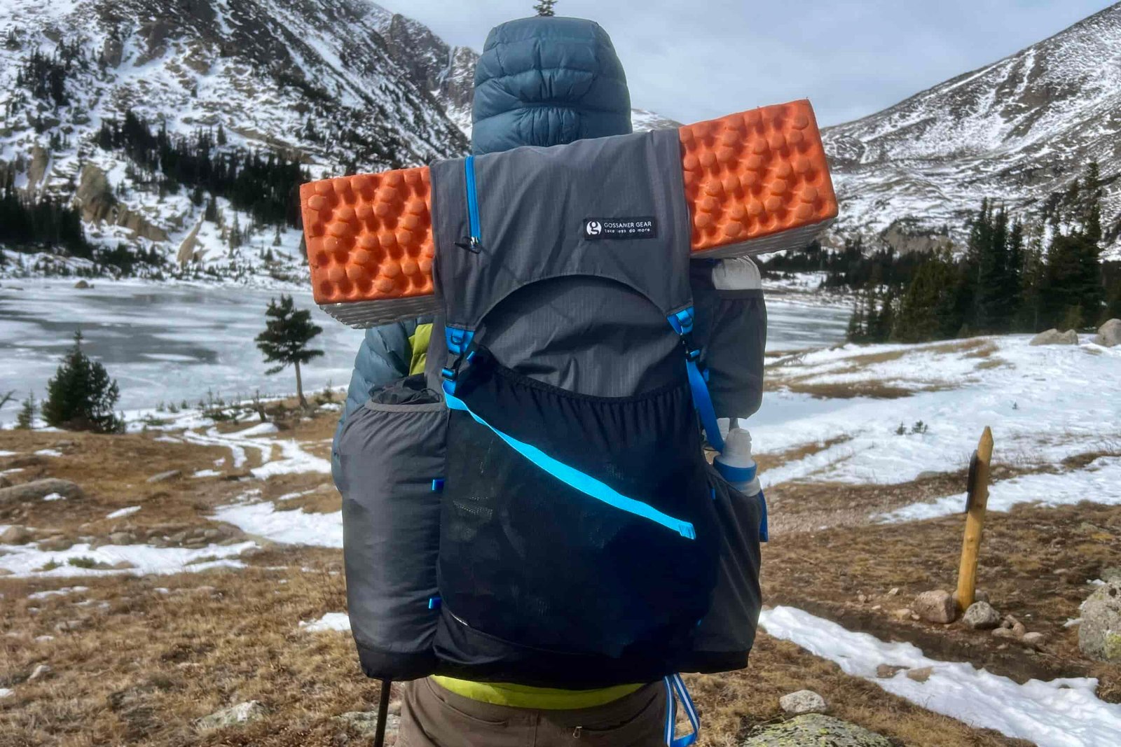 A hiker faces away from the camera overlooking a frozen alpine lake with snow mountains in the background while wearing a fully loaded backpacking backpack.