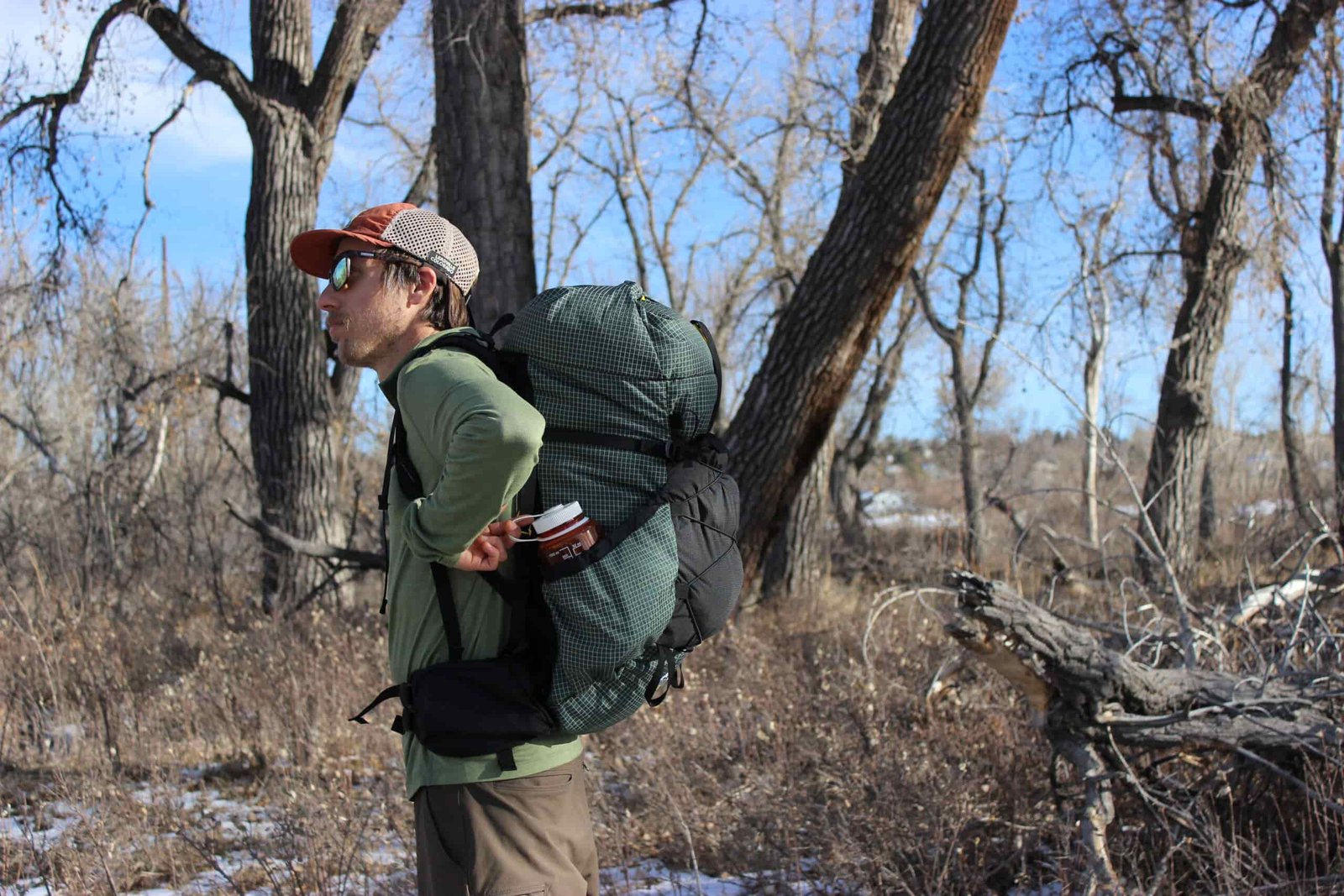 A hiker wearing a green backpack and reaching into the side pockets with trees and blue sky in the background.