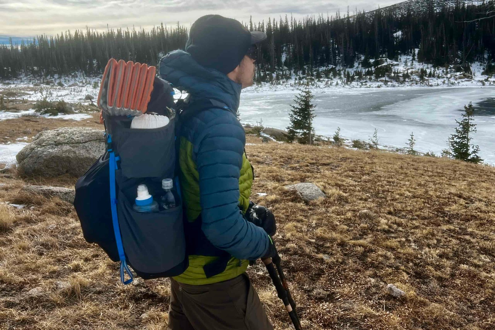 A hiker looks over a frozen lake while wearing a fully packed Mariposa 60 pack. Evergreen trees are in the background.