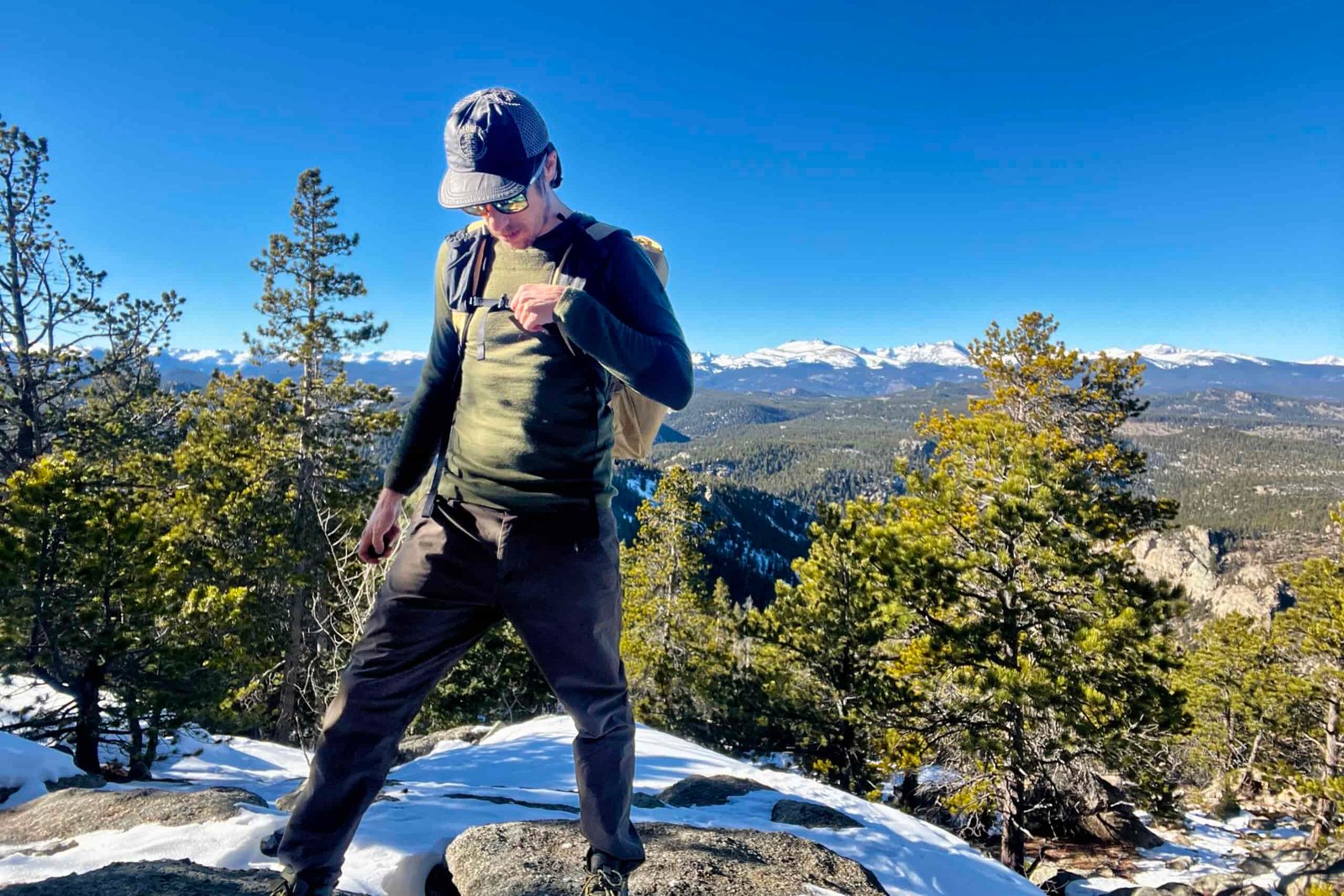 A hiker tightens the sternum strap on his backback while wearing the Smartwool Intraknit. Sky, mountains, and trees in the background.