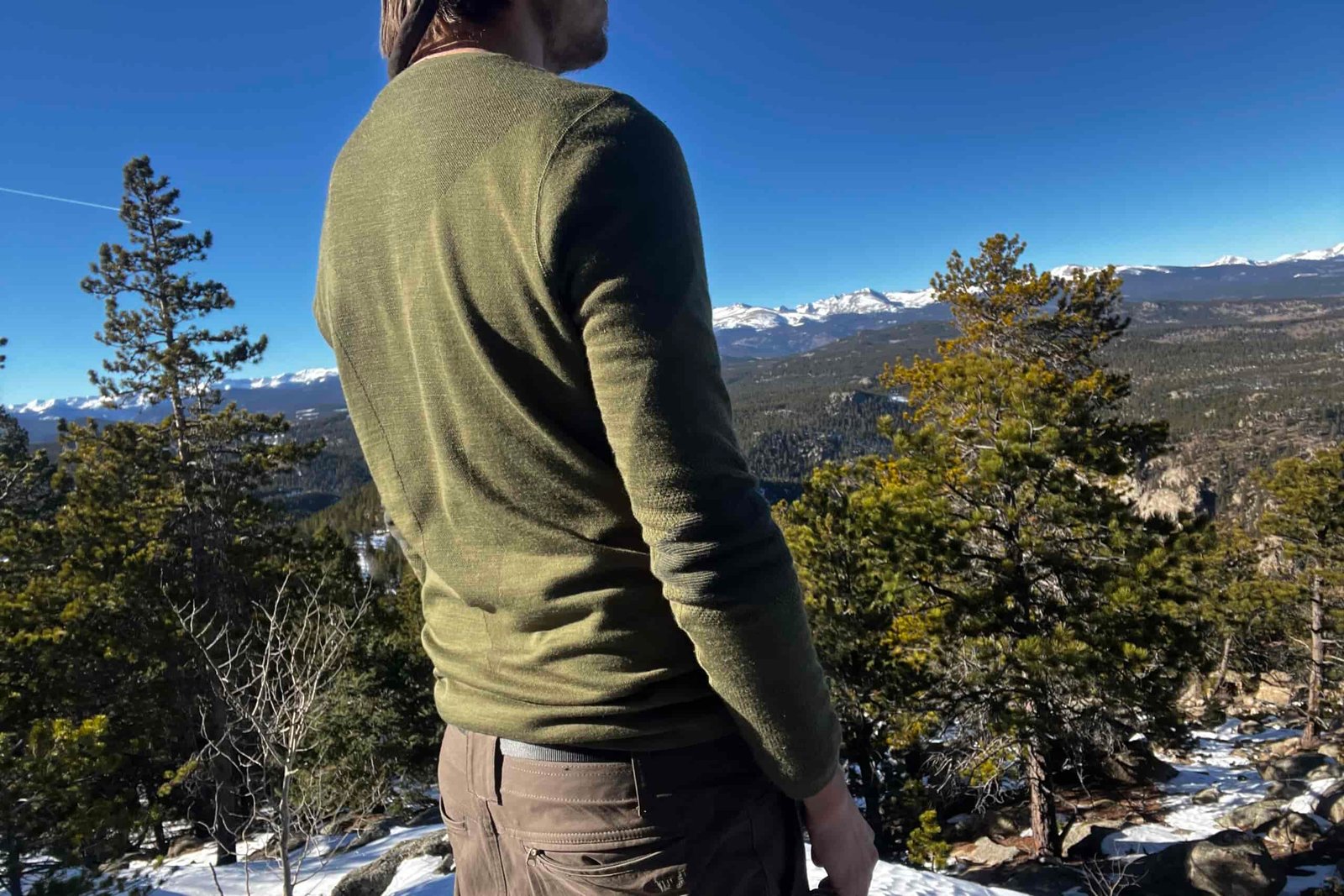 A hiker faces diagonally away from the camera wearing the Smartwool Intraknit base layer. Mountains, trees, and sky in the background.