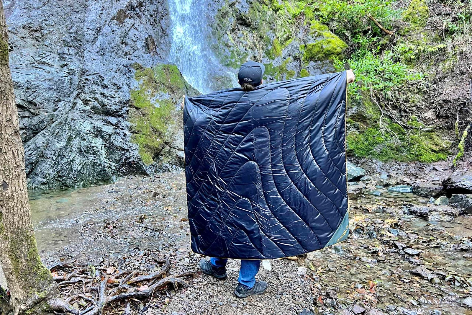 Man standing before a waterfall with the Rumpl Original Puffy Blanket around his back.