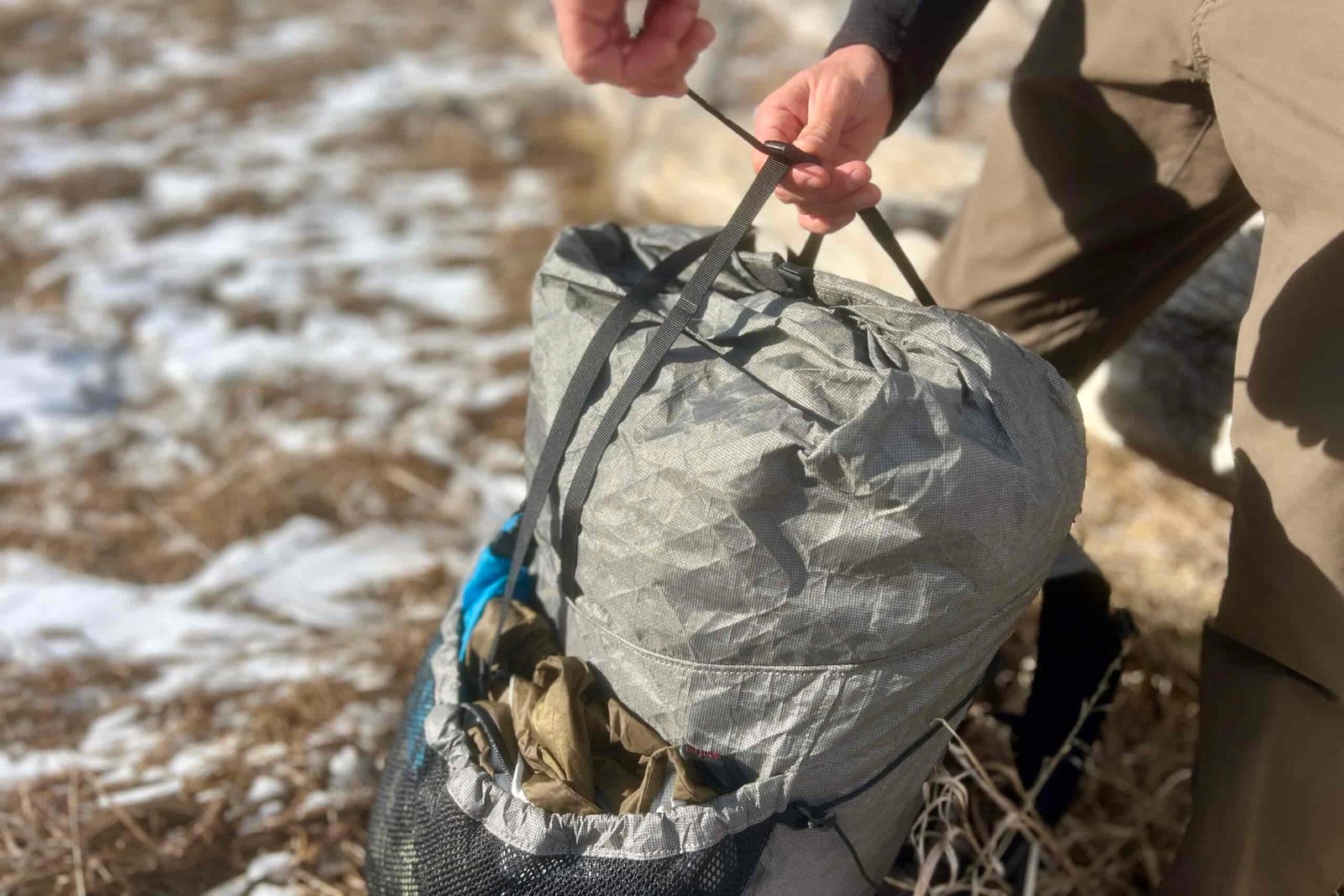 A hiker pulls the top strap of a grey backpacking backpack with snow in the background.