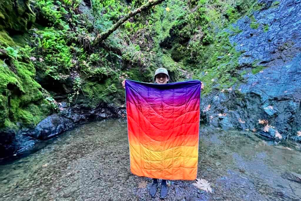 Person holding up the Rumpl NanoLoft Puffy Blanket with a mossy wooded background, to compare blanket size to the person’s height.
