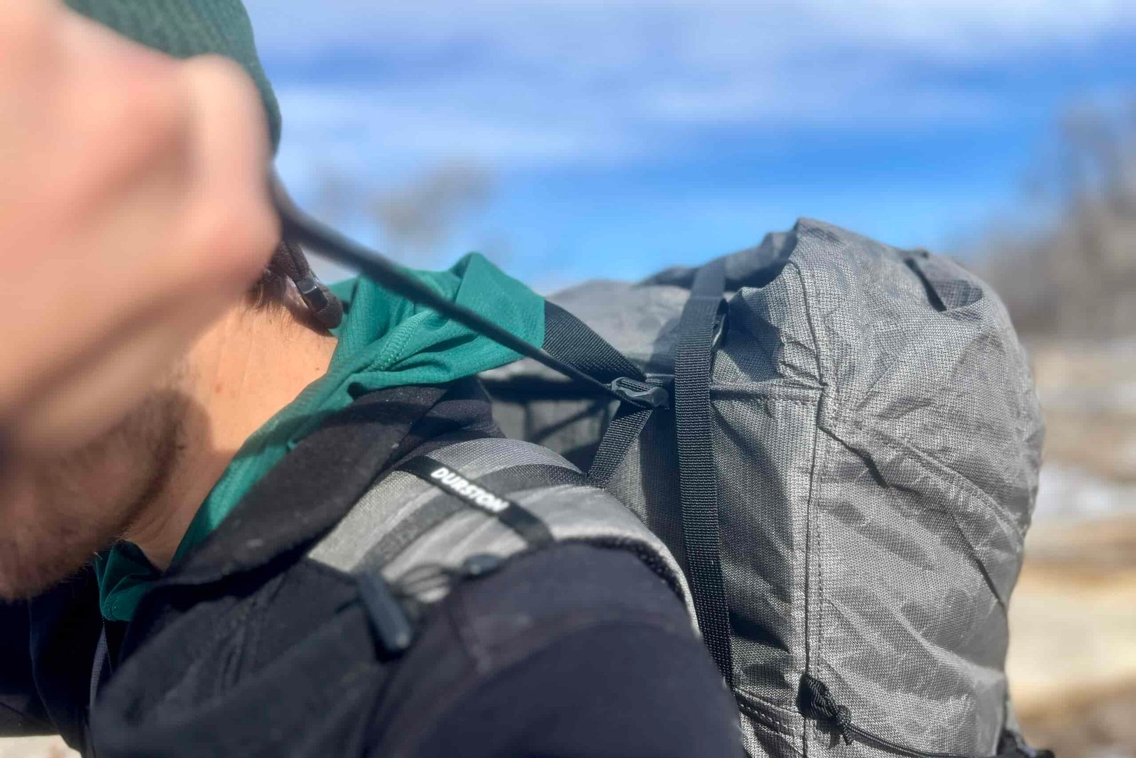 Detail shot of the backpack's load lifters with the wearer pulling the webbing. The image demonstrates how the shoulder straps are constructed and attached to the main pack body, with black webbing visible for adjustments.