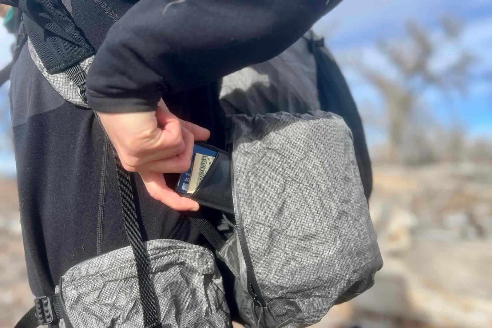 Close-up shot of someone accessing a side zipper pocket on a gray ultralight backpack. Their hand is pulling out a wallet from the zippered pocket.