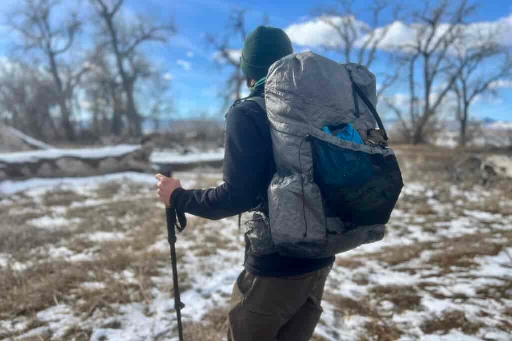 Side view of a hiker wearing a gray ultralight backpack in a snowy winter landscape. The hiker is wearing a black base layer and green beanie while using a trekking pole. The backpack appears to have mesh side pockets and compression straps, with bare winter trees and patches of snow visible in the background.